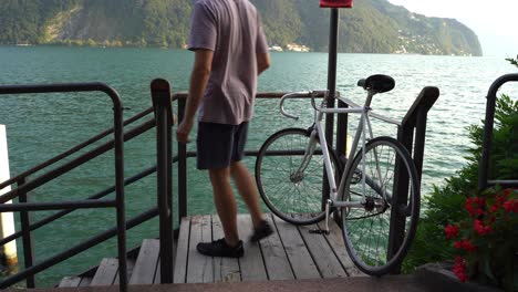 Der-Typ-Erreicht-Sein-Ziel-Und-Lässt-Sein-Fahrrad-Offen-Auf-Dem-Deck-In-Der-Nähe-Eines-Sees-Stehen,-Um-Abenteuer-Zu-Erleben