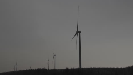 Line-of-wind-turbines-on-woodland-covered-hills-into-the-distance,-spinning-with-sky-background