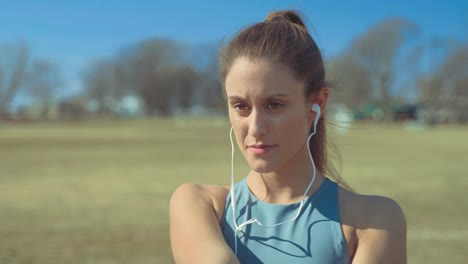 Portrait-Of-Young-Woman-Putting-On-Earphones-To-Listen-To-Music,-Then-Begins-Stretching-To-Warm-Up-For-Her-Outdoor-Workout-On-Sunny-Day