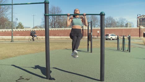 Fit-Caucasian-Girl-Working-Out-With-Pull-Up-Exercises,-Strength-Training-At-Outdoor-Gym-On-Sunny-Summer-Day---Wide-Shot