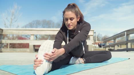 Young-Woman-Stretching-Leg,-Sitting-On-Yoga-Mat-On-Wooden-Pier,-Wearing-Black-Sweats,-On-Sunny-Day-Before-Exercising