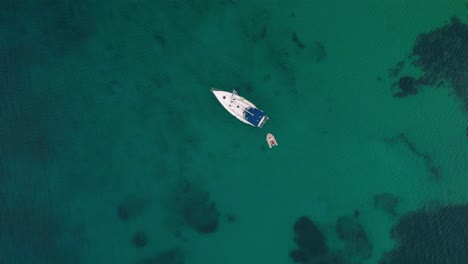 Aerial-view-rising-above-a-sailboat-on-the-turquoise,-Mediterranean-sea,-on-the-coast-of-Mykonos,-dark,-cloudy-day,-in-Greece---screwdriver,-drone-shot
