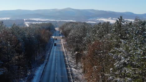 Straße-Mit-Fahrzeugen,-Die-Mitten-In-Einem-Winterwald-Mit-Schneebedeckten-Nadelbäumen-Fahren---Luftdrohnenrückzug