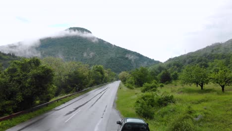 Car-Driving-On-Wet-Road-In-Rural-Area-With-Foggy-Hill-In-Background---aerial