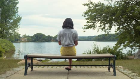 Female-Student-Reading-Book-On-Park-Bench-By-Calm,-Peaceful-Lake,-Wide-Shot,-Filmed-From-Behind