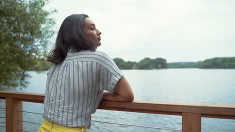 Beautiful-Latina-Woman-Relaxed,-Leaning-On-Dock-Railing-By-Calm-Lake,-Closes-Her-Eyes-To-Take-Deep-Breath-Inhale-and-Exhale,-Slow-Motion-Shot