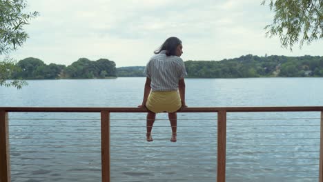 Young-Woman-Sitting-Alone-In-Peace-On-Dock-Railing-By-Pond-Enjoying-The-Views-Of-The-Calm-Water