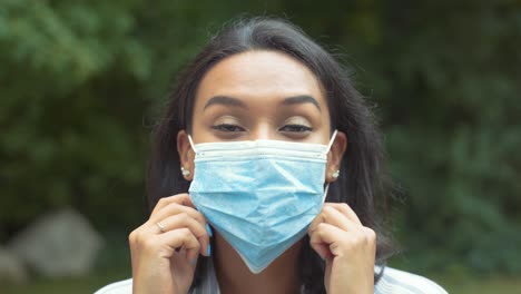Portrait-Of-Pretty-Latin-Woman-Putting-On-Medical-Face-Mask-With-Both-Hands-While-Tucking-Hair-Behind-Ears,-Outside-At-Park