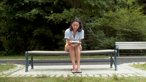 Young-Hispanic-Woman-Reading-Book,-Sitting-With-Legs-Crossed-On-Bench-At-Park,-Wide-Shot