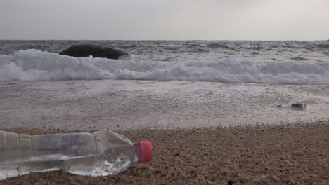 Littered-plastic-drinking-bottle-left-on-a-beach-near-waves,-steady-low-angle-shot