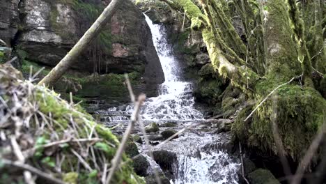 4K,-long-shot-with-foreground-elements,-small-waterfall-flowing-over-rocks-in-forest,-South-Wales,-UK