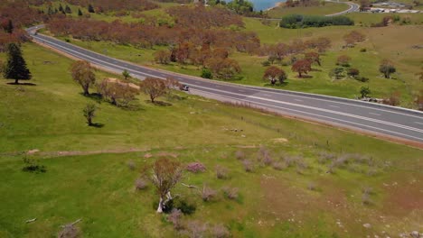 Aerial-view-over-a-road,-dry-trees-and-meadows,-towards-lake-Jindabyne,-in-Australia---tilt-up,-drone-shot