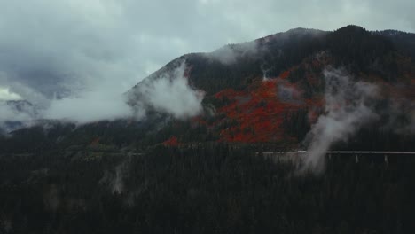Washington-State---Drone-aerial-static-of-a-misty-pine-forest-and-mountains-with-a-highway,-trucks,-and-cars,-and-red-fall-colours-near-Franklin-Falls-and-Snowqualmie
