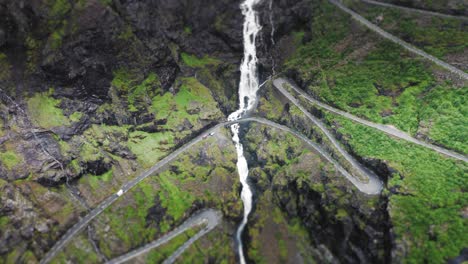An-spectacular-aerial-view-on-Stigfossen-waterfall-A-massive-water-flow-is-falling-down-the-mountain-overgrown-with-green-grass,-cut-in-with-a-two-lane-serpentine-road,-and-a-bridge-over-the-waterfall