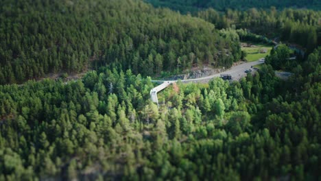 A-narrow-asphalt-road-winding-uphill,-leading-to-the-Stegastein-viewpoint-Dense-pine-forest-covering-the-mountainside-The-viewpoint-platform-hanging-from-the-cliff-overlooking-the-fjord-and-the-valley