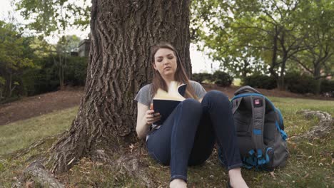 Female-College-Student-Takes-Notebook-Out-Of-Backpack-To-Write-In-Journal-Under-A-Tree-At-Campus-Park
