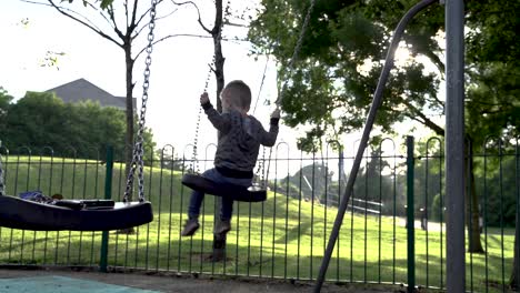 Three-Year-Old-Boy-being-pushed-on-swing-in-late-afternoon-summer-sun-setting-behind-him,-in-playground,-UK