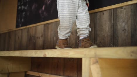 Toddler-boy-slowly-walks-right-along-wooden-bench-in-hut,-mid-shot-feet