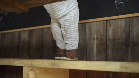 Toddler-boy-stands-on-wooden-bench-and-leans-on-wooden-table-in-hut