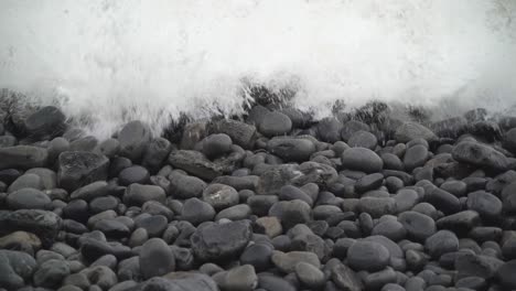 Close-up-on-pebbles-as-white-winter-waves-crash-in-on-beach-at-Dunraven-Bay,-South-Wales