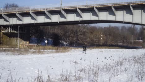 Man-ride-a-bike-on-snow-during-winter-under-bridge-with-cars-and-tram