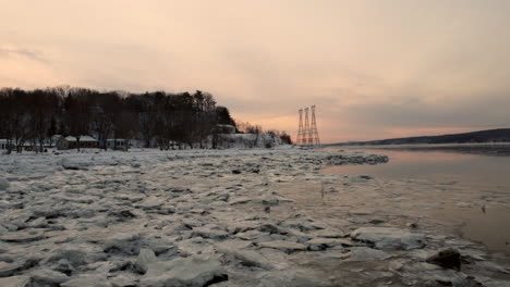 Aerial---Flying-over-the-icy-shore-of-the-St-Lawrence-river,-towards-power-transmission-lines,-at-sunset