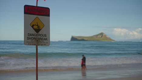 A-slow-pan-shot-of-the-dangerous-shorebreak-warning-sign-with-a-parent-playing-with-their-kids