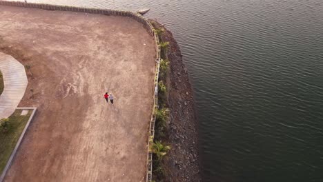 4k-aerial-view-video-of-a-couple-walking-with-a-Shih-Tzu-Small-Dog-Along-the-Shore-of-a-Beautiful-Sceinic-Lake-on-Vacation-or-holiday-During-Summer-near-Pune,-India