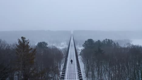 Cinematic-ascending-aerial-shot-of-a-man-walking-along-a-very-long-railway-bridge,-in-a-snowy-environment