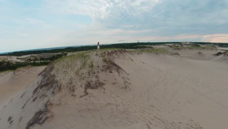 Drone-zoom-in-follow-up-shot-of-the-amazing-barren-desert-landscape-and-a-girl-running-on-the-high-sand-dunes-and-trails-in-the-Dune-Shacks-Trail-in-Provincetown,-Cape-Cod,-Massachusetts