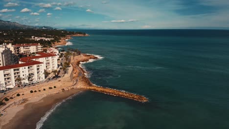Zoom-out-of-the-coastal-promenade-of-Ametlla-de-Mar-in-Tarragona,-Spain-with-waves-crashing-against-the-shore-on-a-sunny-day---Aerial-view