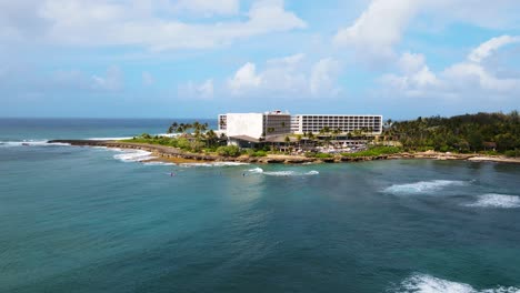 Cinematic-drone-shot-of-the-blue-waters-and-building-in-the-background-near-the-turtle-bay-coastline,-Oahu-Hawaii