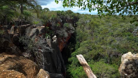 Tilting-down-shot-looking-out-to-a-hole-in-the-ground-created-from-the-Lapa-Doce-cave-with-a-jungle-inside-and-colorful-cliffs-in-the-Chapada-Diamantina-National-Park-in-Bahia,-northeastern-Brazil