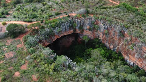 Aerial-drone-wide-shot-of-the-large-Lapa-Doce-cave-entrance-of-colorful-rocks-with-a-self-contained-rainforest-below-in-the-Chapada-Diamantina-National-Park-in-Bahia,-northeastern-Brazil