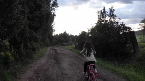 Tracking-shot-of-a-young-girl-riding-her-bike-down-an-uneven-dirt-road