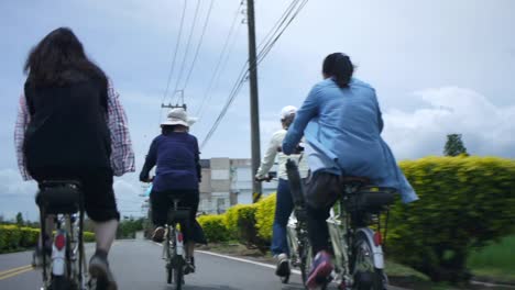 Grupo-De-Mujeres-Ciclistas-Que-Viajan-Por-Una-Carretera-Rural-Junto-A-Las-Líneas-Eléctricas,-Filmadas-Desde-Atrás-Siguiendo-A-Los-Ciclistas