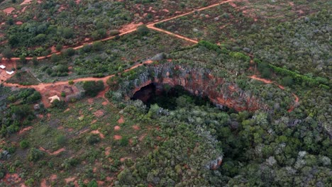 Aerial-drone-rotating-wide-shot-of-the-large-Lapa-Doce-cave-entrance-of-colorful-rocks-with-a-self-contained-rainforest-below-in-the-Chapada-Diamantina-National-Park-in-Bahia,-northeastern-Brazil