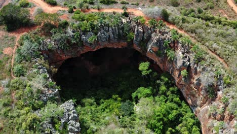 Aerial-drone-rotating-medium-shot-of-the-large-Lapa-Doce-cave-entrance-of-colorful-rocks-with-a-self-contained-rainforest-below-in-the-Chapada-Diamantina-National-Park-in-Bahia,-northeastern-Brazil