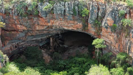 Aerial-drone-medium-shot-of-the-large-Lapa-Doce-cave-entrance-of-colorful-rocks-with-a-self-contained-rainforest-below-in-the-Chapada-Diamantina-National-Park-in-Bahia,-northeastern-Brazil