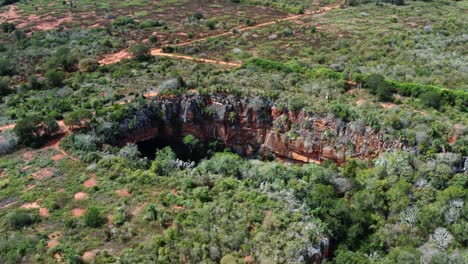 Aerial-drone-wide-shot-of-the-large-Lapa-Doce-cave-entrance-of-colorful-rocks-with-a-self-contained-rainforest-below-in-the-Chapada-Diamantina-National-Park-in-Bahia,-northeastern-Brazil