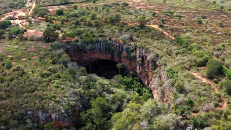 Aerial-drone-trucking-left-shot-of-the-large-Lapa-Doce-cave-entrance-of-colorful-rocks-with-a-self-contained-rainforest-below-in-the-Chapada-Diamantina-National-Park-in-Bahia,-northeastern-Brazil