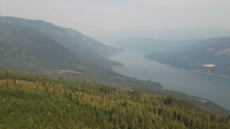 Schöne-Luftaufnahme-Des-Waldes-Auf-Einem-Hügel-Mit-Nebligen-Bergen-Und-See-Als-Hintergrund