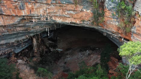 Aerial-drone-medium-dolly-out-shot-of-the-large-Lapa-Doce-cave-entrance-of-colorful-rocks-with-a-self-contained-rainforest-below-in-the-Chapada-Diamantina-National-Park-in-Bahia,-northeastern-Brazil