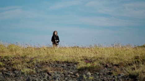 young-girl-walking-in-tall-grass-field,-coming-toward-the-camera-on-a-sunny-day-with-some-clouds-in-the-background