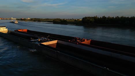 Aerial-Orbital-View-of-Nautical-Traffic,-Long-Cargo-Barge-Inland-Waterway-Vessel-Navigating-Along-River-Canal-in-Zwijndrecht-Netherlands-at-Sunset,-Coastline-Navigation-and-Transportation