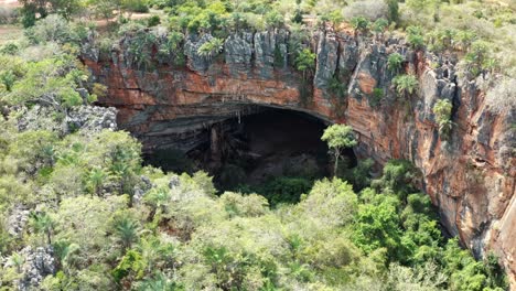 Aerial-drone-dolly-out-shot-of-the-large-Lapa-Doce-cave-entrance-of-colorful-rocks-with-a-self-contained-rainforest-below-in-the-Chapada-Diamantina-National-Park-in-Bahia,-northeastern-Brazil