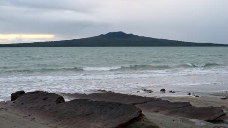 Beautiful-Rangitoto-island,-a-dormant-volcano-visible-from-a-rocky-beach-in-north-Auckland,-New-Zealand