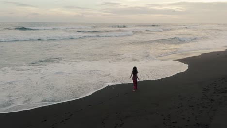 Woman-In-Red-Dress-Walking-On-The-Black-Sand-Beach-In-El-Paredon,-Guatemala-At-Sunset---aerial-drone-shot