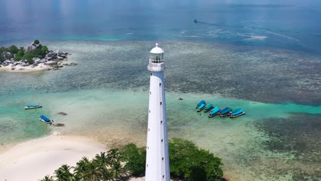 Zoom-Aéreo-Fuera-Del-Faro-Blanco-En-La-Isla-Lengkuas-Con-Barcos-Turísticos-Anclados-En-Aguas-Tropicales-De-Belitung