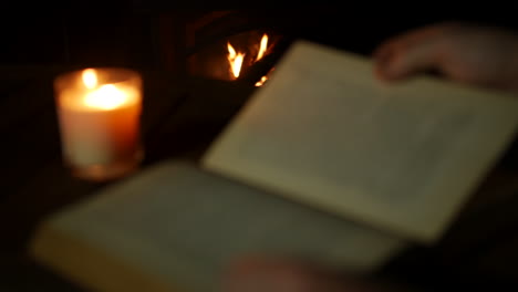 Ein-Feuer,-Das-In-Einem-Kamin-Brennt,-Während-Ein-Mann-Ein-Altes-Antikes-Buch-Liest,-Das-Von-Kerzenlicht-Beleuchtet-Wird